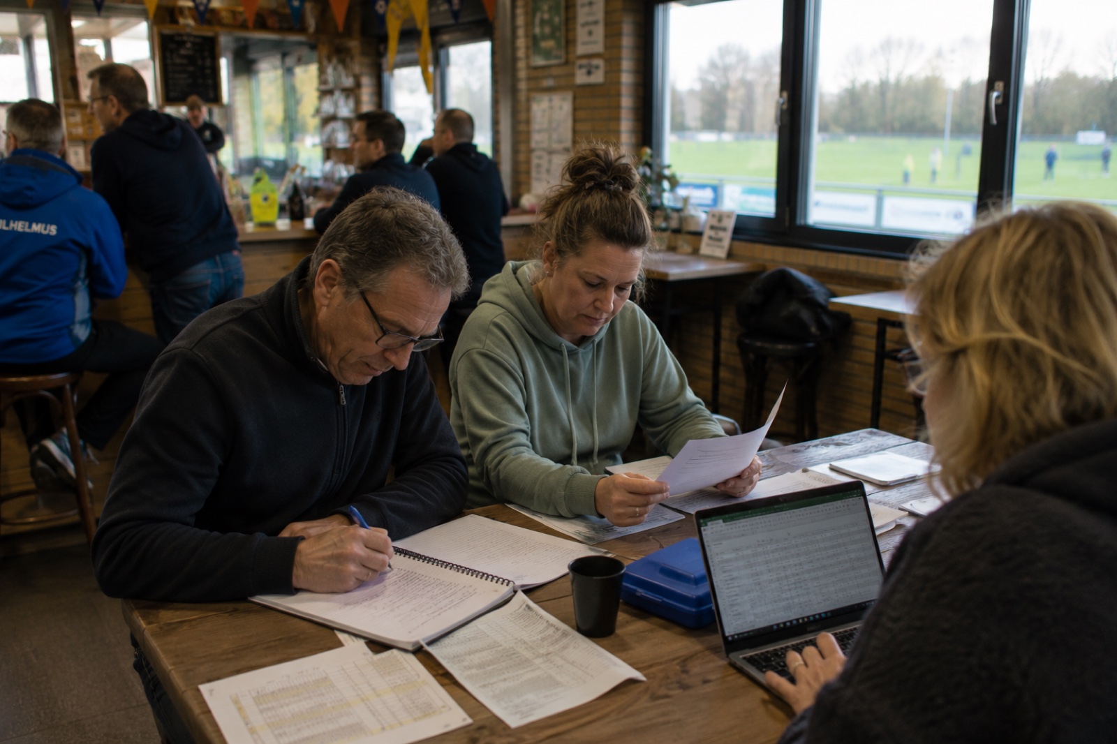 Drie mensen aan een tafel vol papieren en een laptop met spreadsheet in een kantine